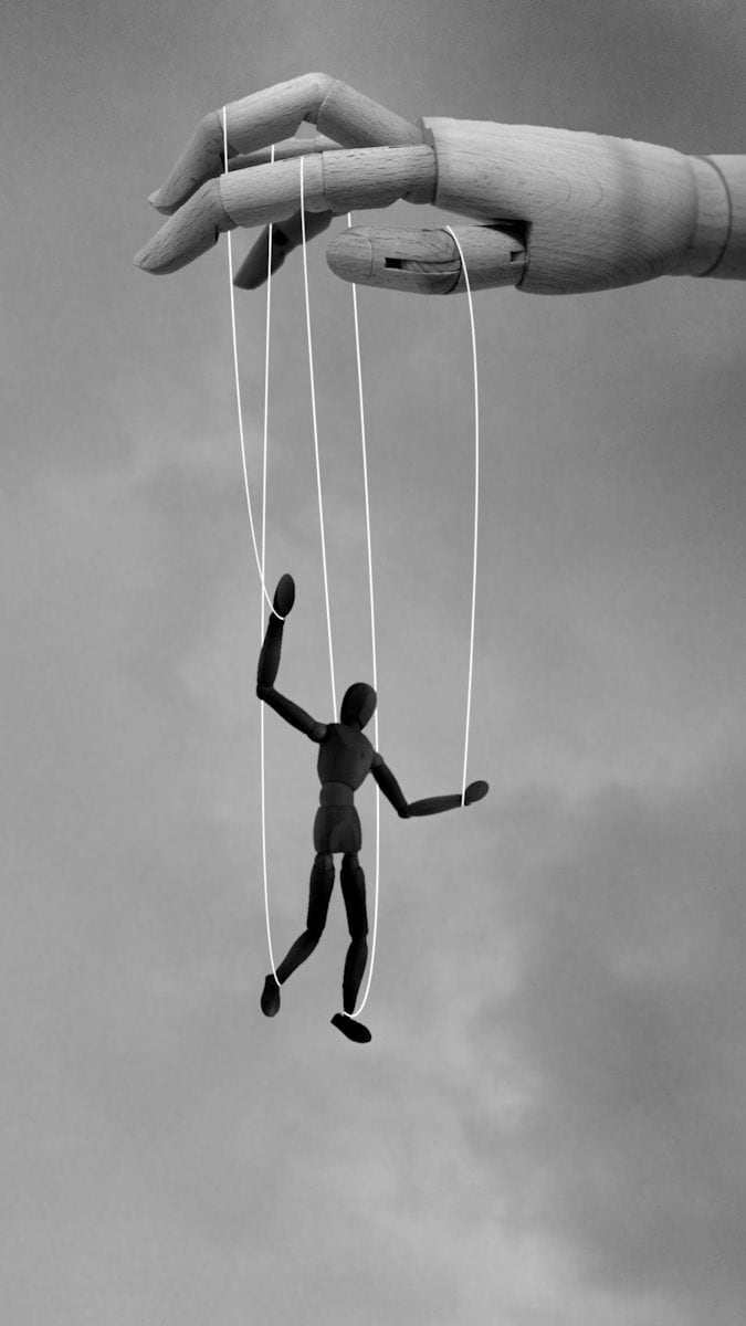A large wooden hand manipulating a small black mannequin with white strings