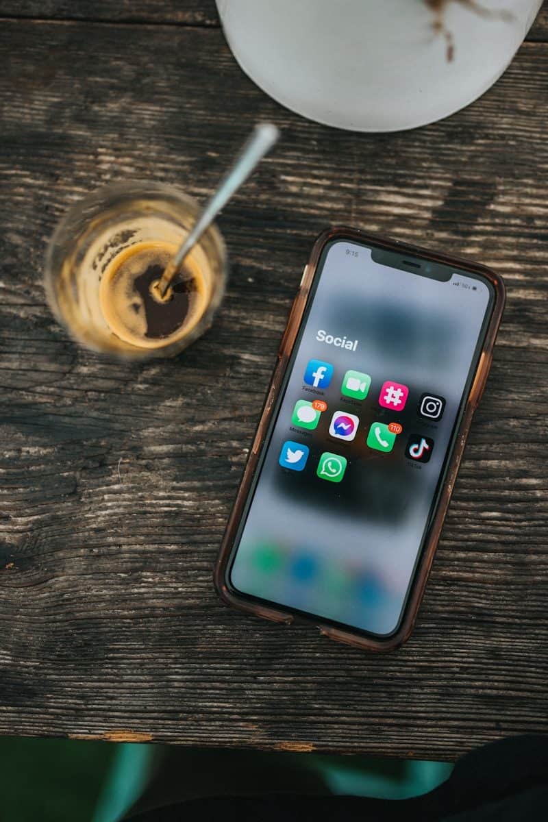 A smartphone displaying social media apps beside a glass of coffee on wood