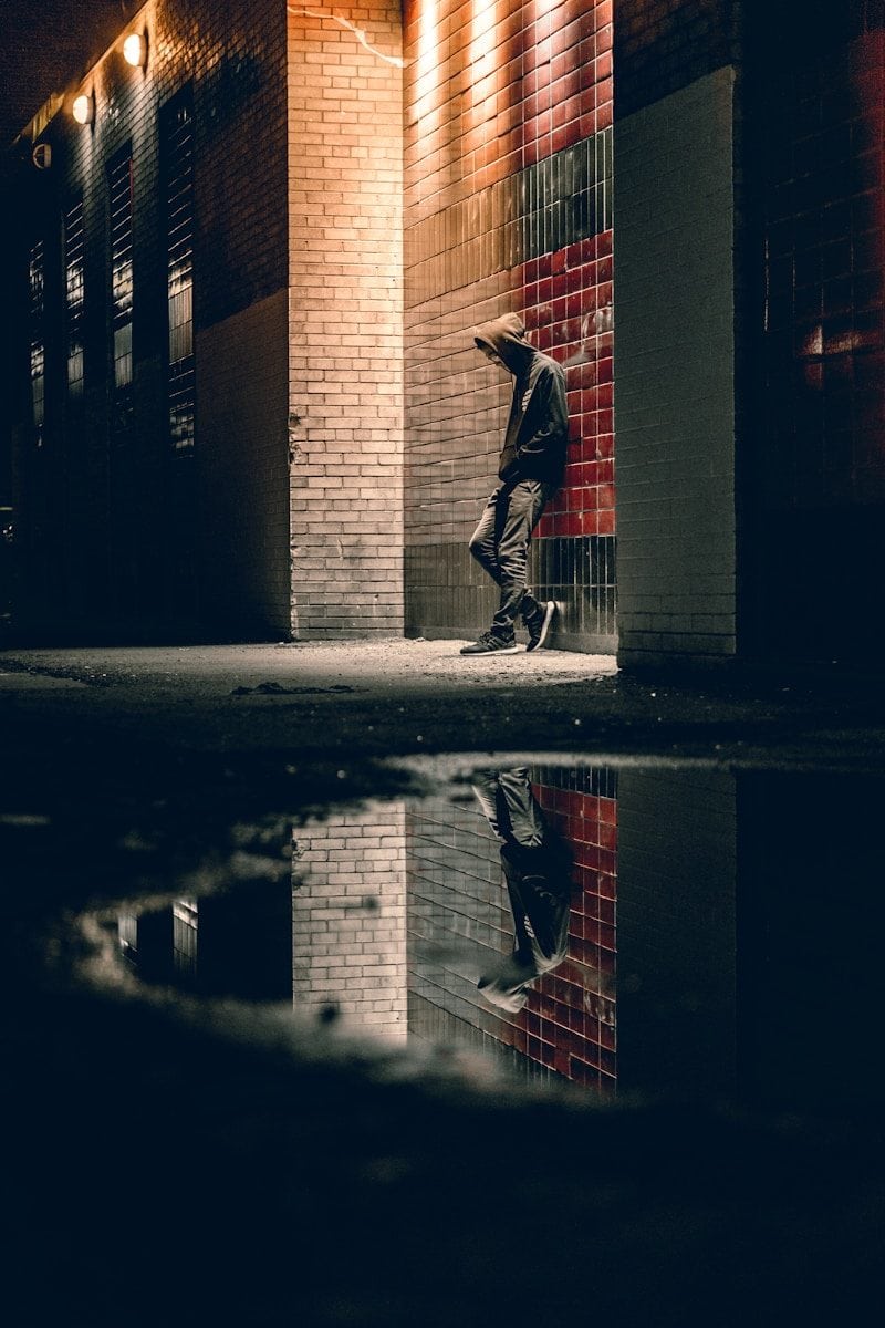 A person in a hooded sweatshirt leaning against a brick wall at night