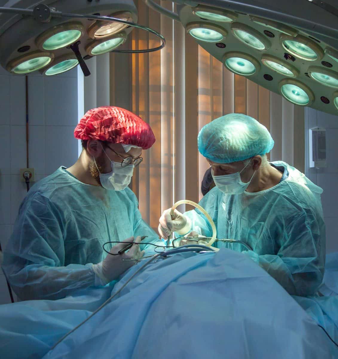 Two surgeons in blue scrubs and masks performing surgery under bright overhead lights