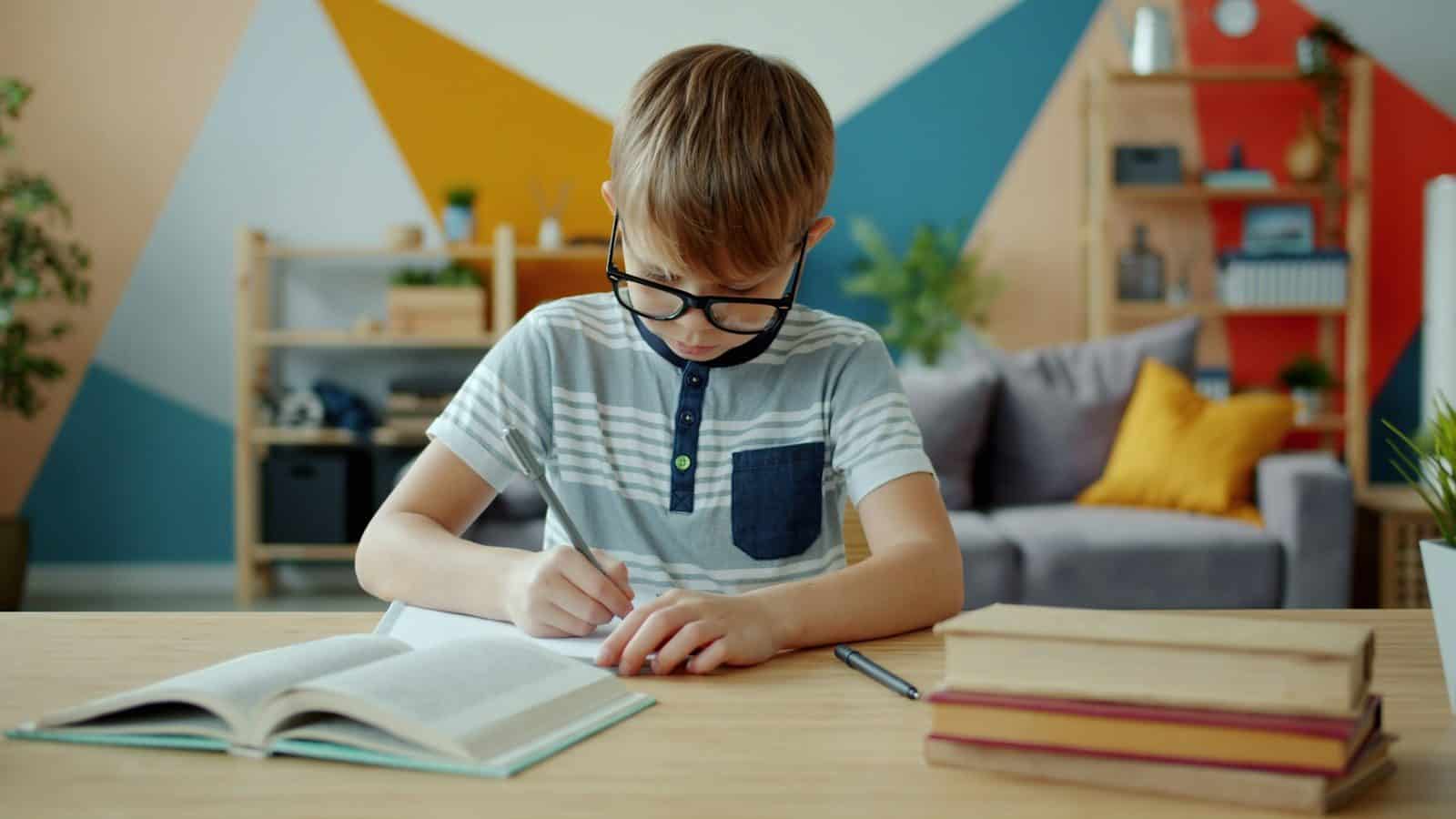 Young boy with glasses writing in a notebook at desk